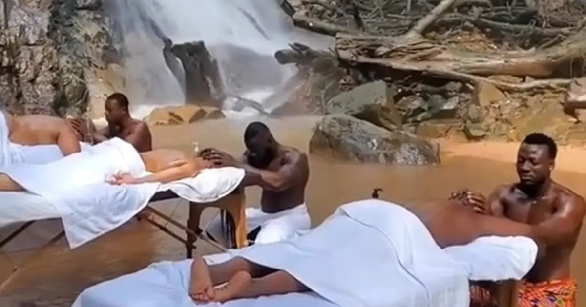 Group of women receiving outdoor massages by shirtless male masseuses in Ghana near a waterfall, surrounded by nature.