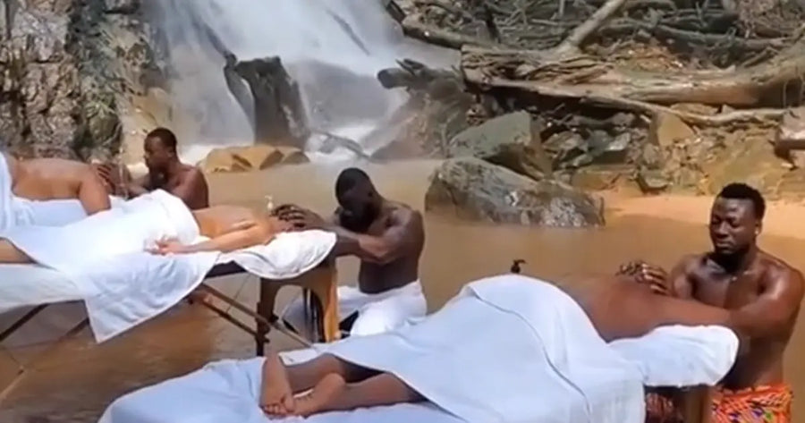 Group of women receiving outdoor massages by shirtless male masseuses in Ghana near a waterfall, surrounded by nature.
